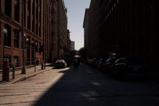 Moody street scene in city with tall buildings, cars, and deep shadows on cobblestones.