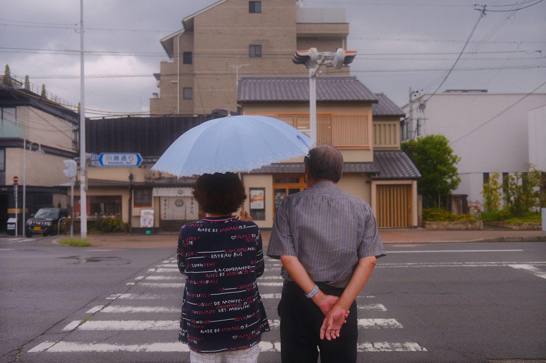 A couple stands at a crosswalk in Kyoto, Japan, under an umbrella on a cloudy day.