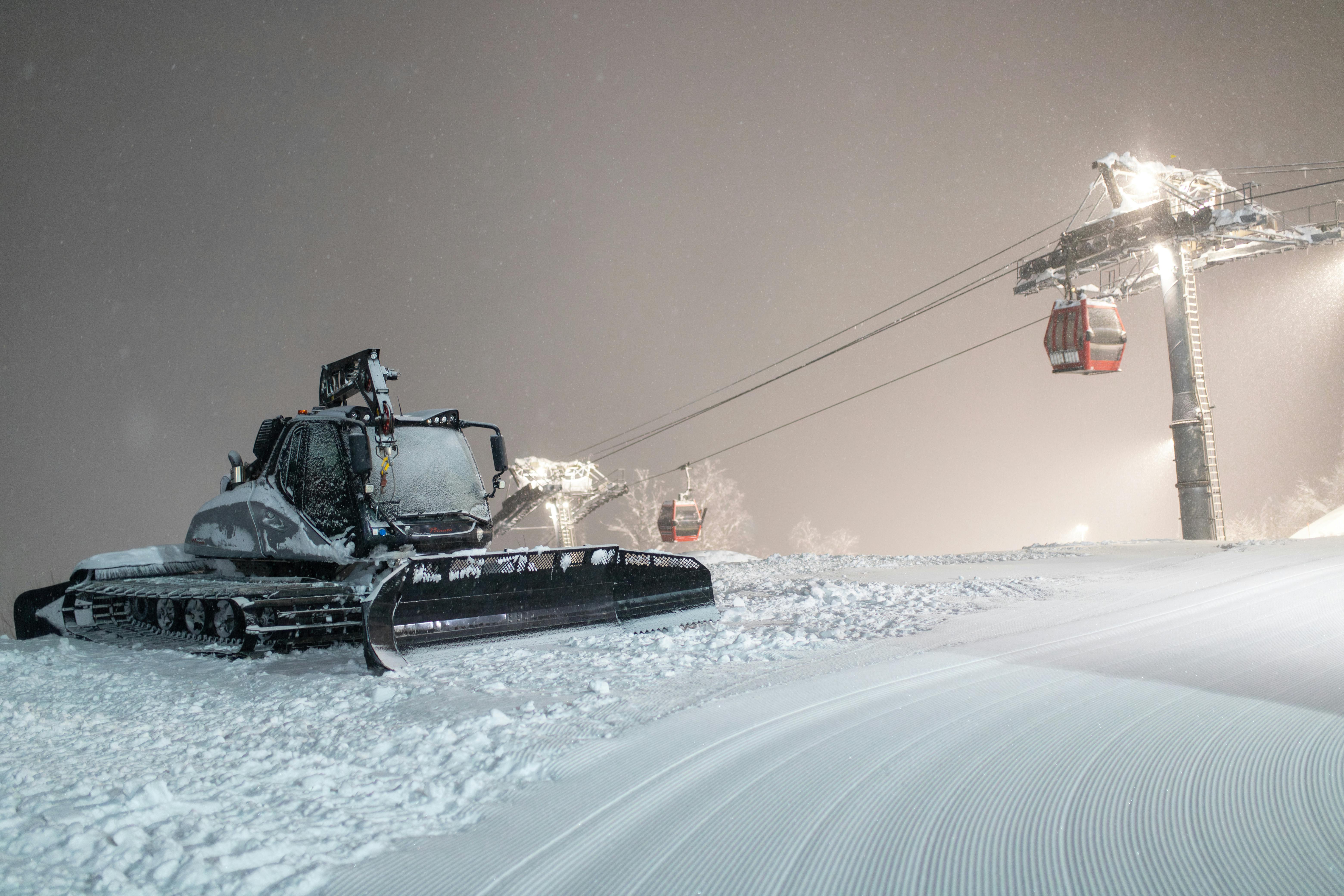 A snowcat working under night lights on a snowy ski slope, with cable cars in the background.