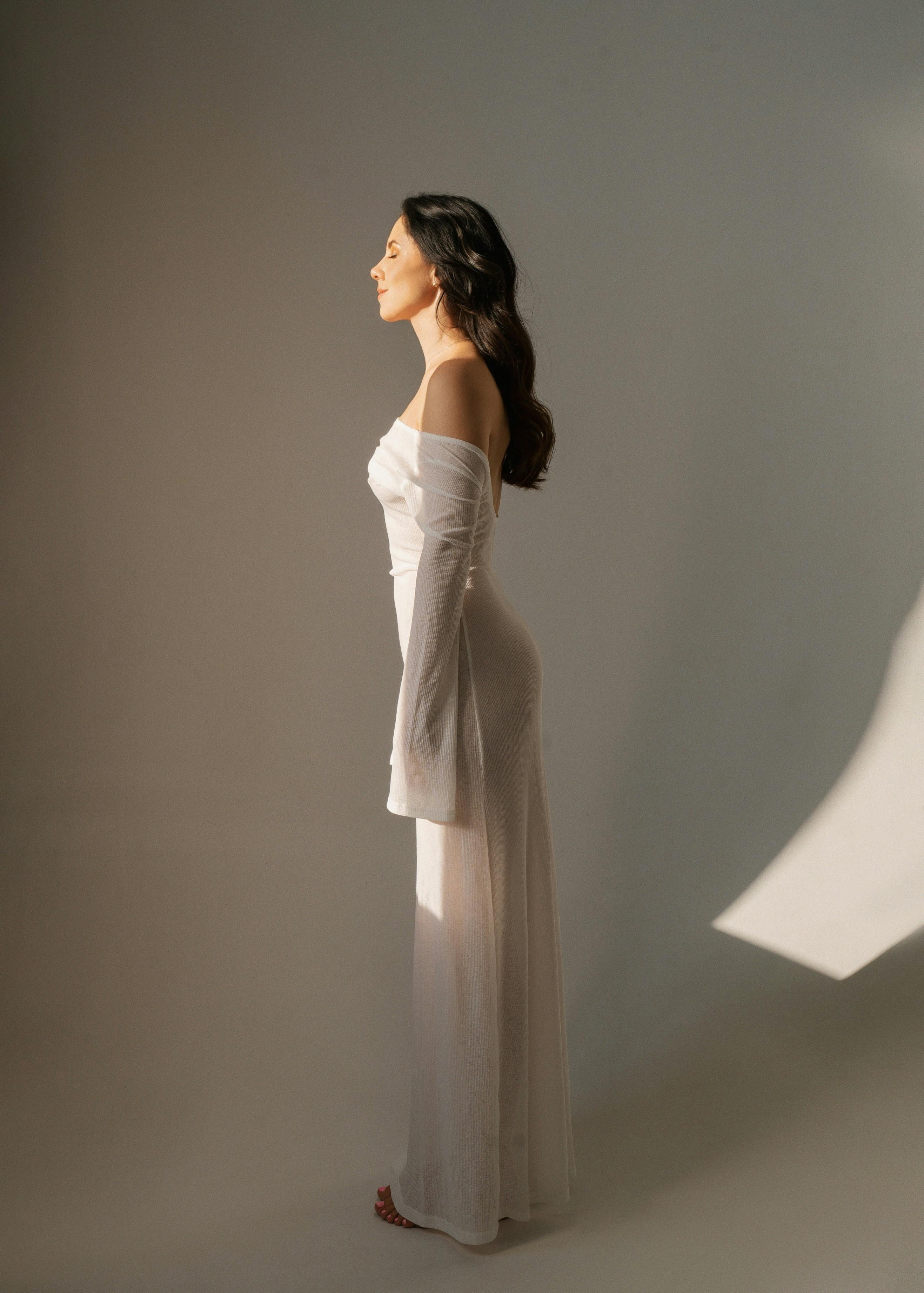 A woman in a white dress poses gracefully in a sunlit studio space.