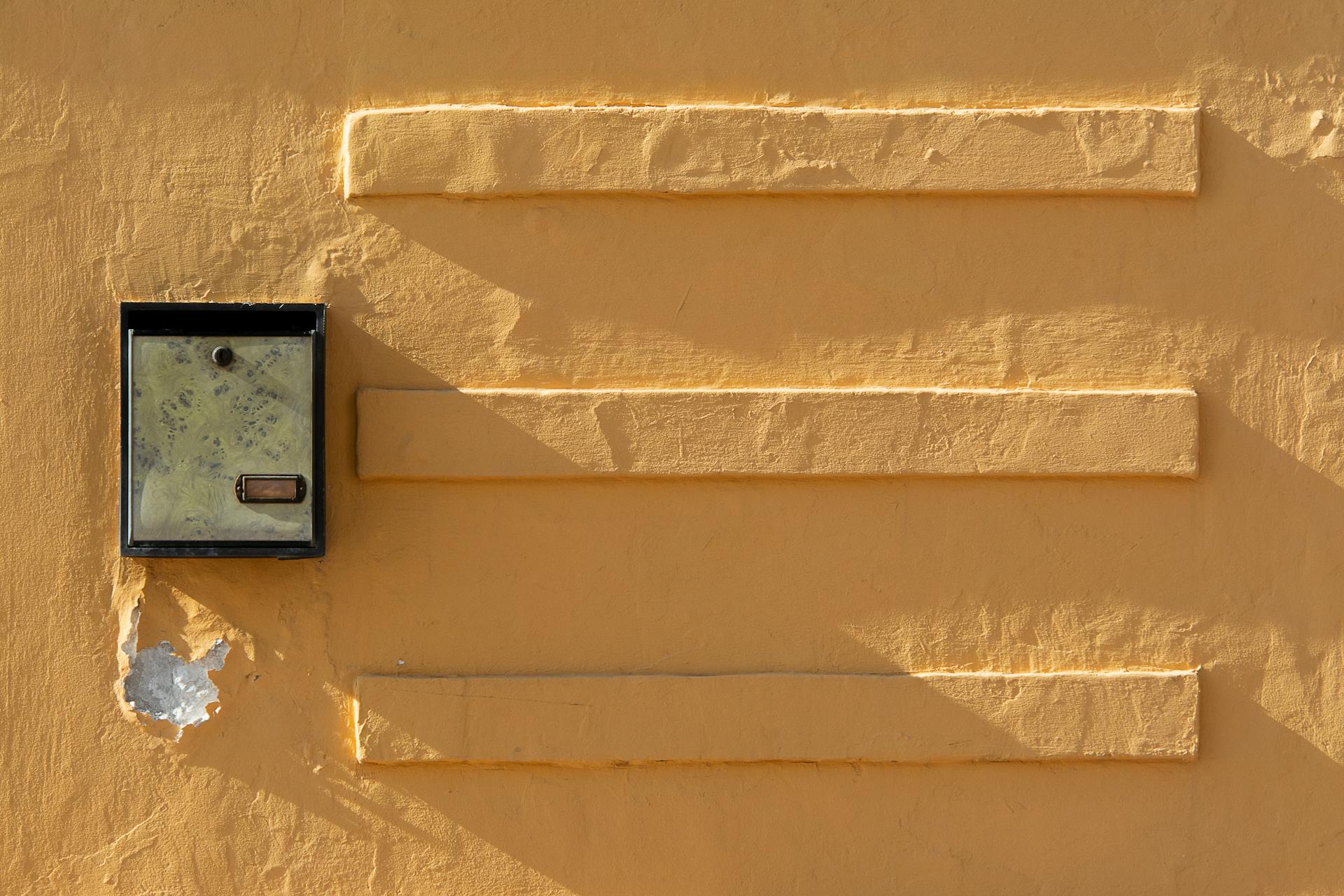 A solitary metal mailbox on a textured yellow wall casting shadows. Ideal for abstract themes.
