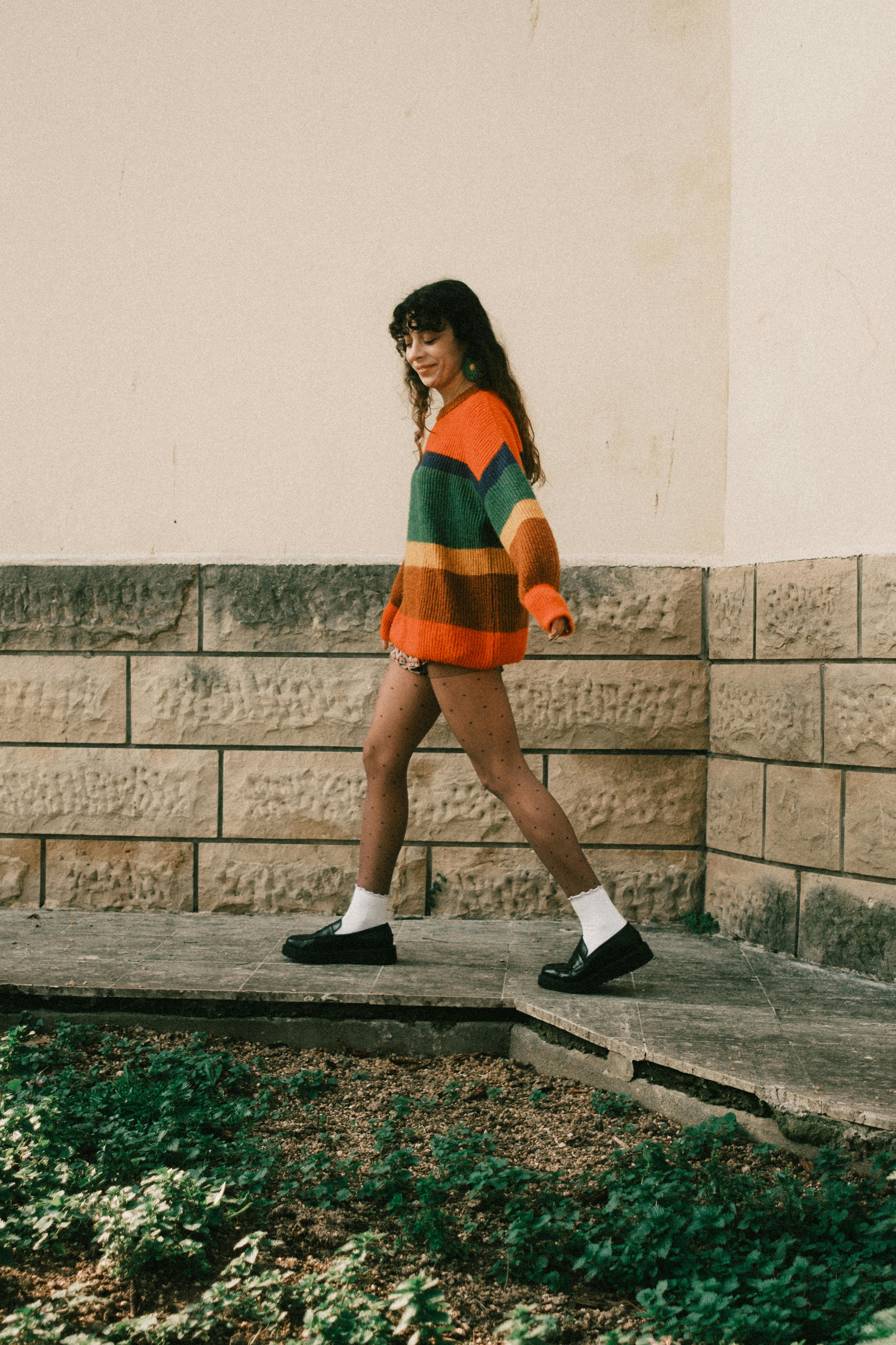 Young woman walks outdoors wearing a colorful striped sweater, exuding casual style.