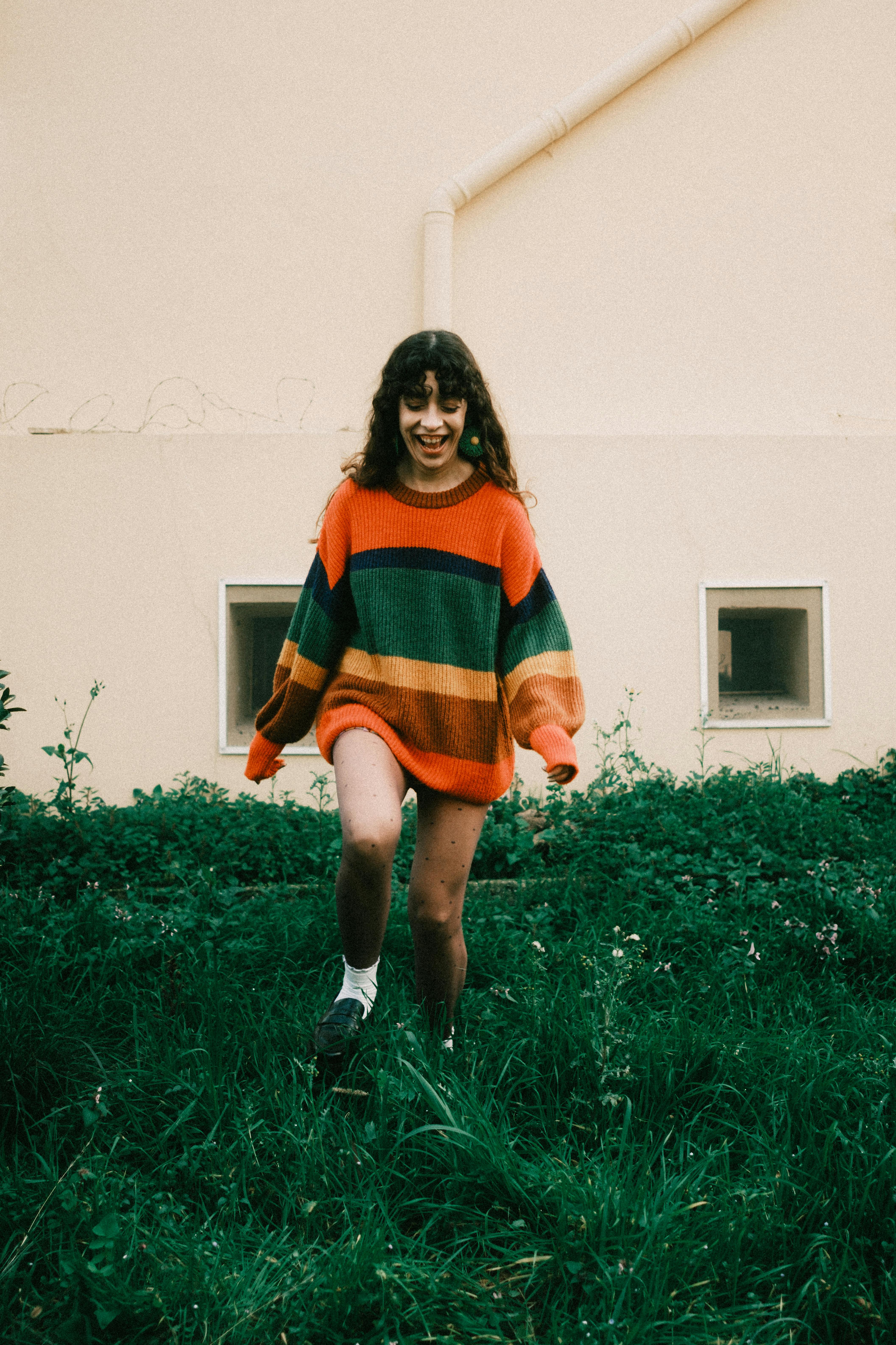 Woman in vibrant striped sweater walking on green grass in front of a yellow wall.