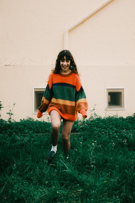 Woman in vibrant striped sweater walking on green grass in front of a yellow wall.