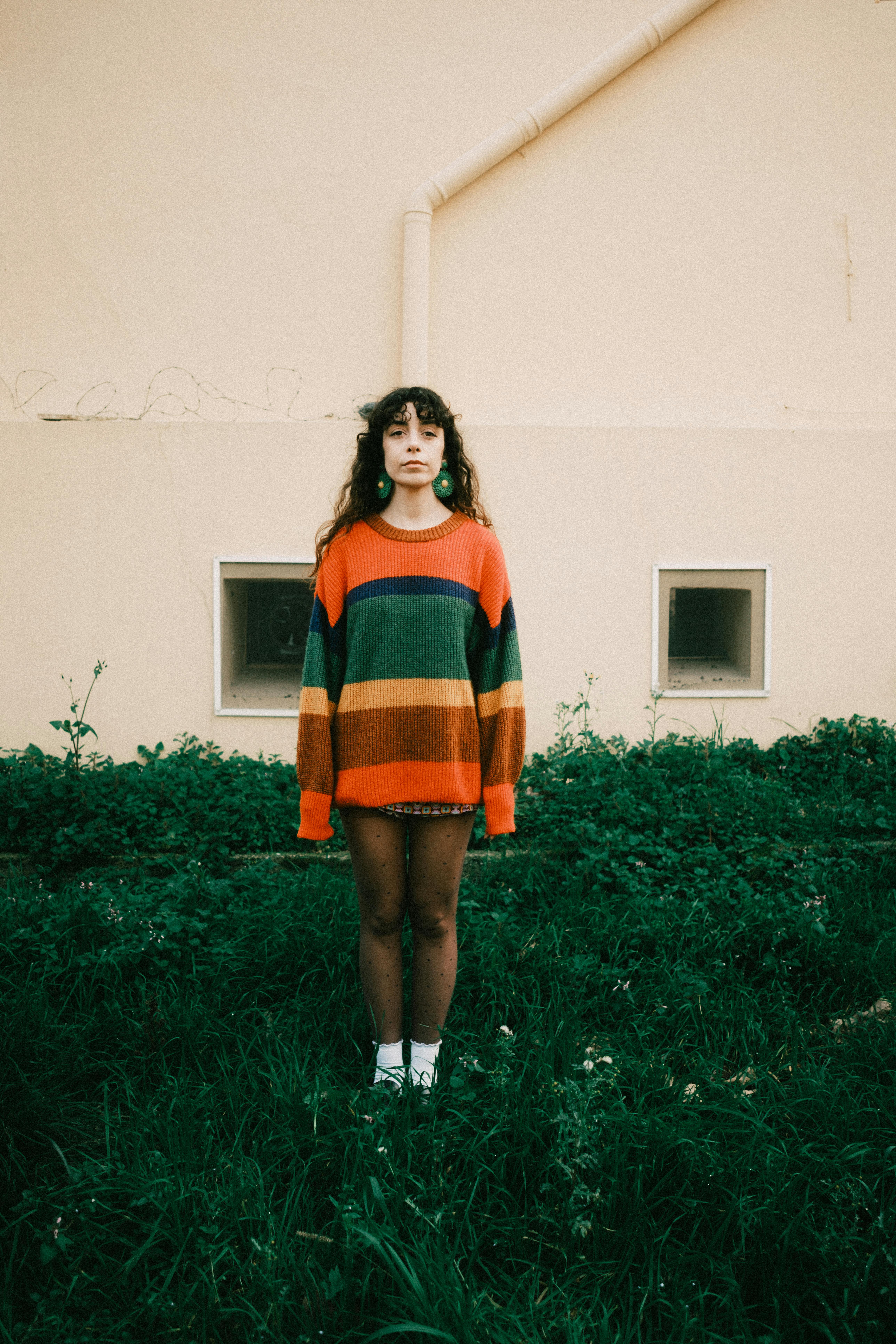 Young woman with curly hair wearing a vibrant striped sweater stands on green grass against a beige wall.