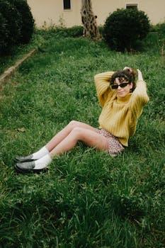 Woman in sunglasses and yellow sweater relaxes on grass outdoors, exuding a stylish, carefree vibe.