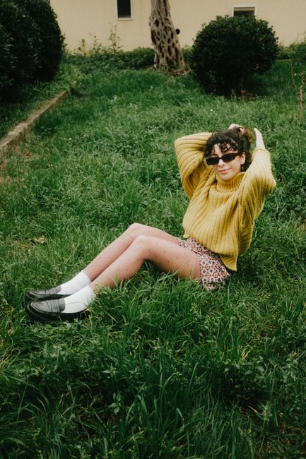 Woman in sunglasses and yellow sweater relaxes on grass outdoors, exuding a stylish, carefree vibe.