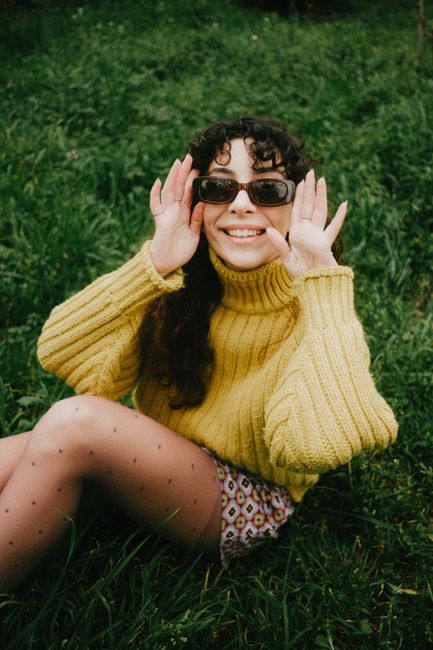 Smiling woman in a yellow sweater sitting in grassy field, adjusting sunglasses.