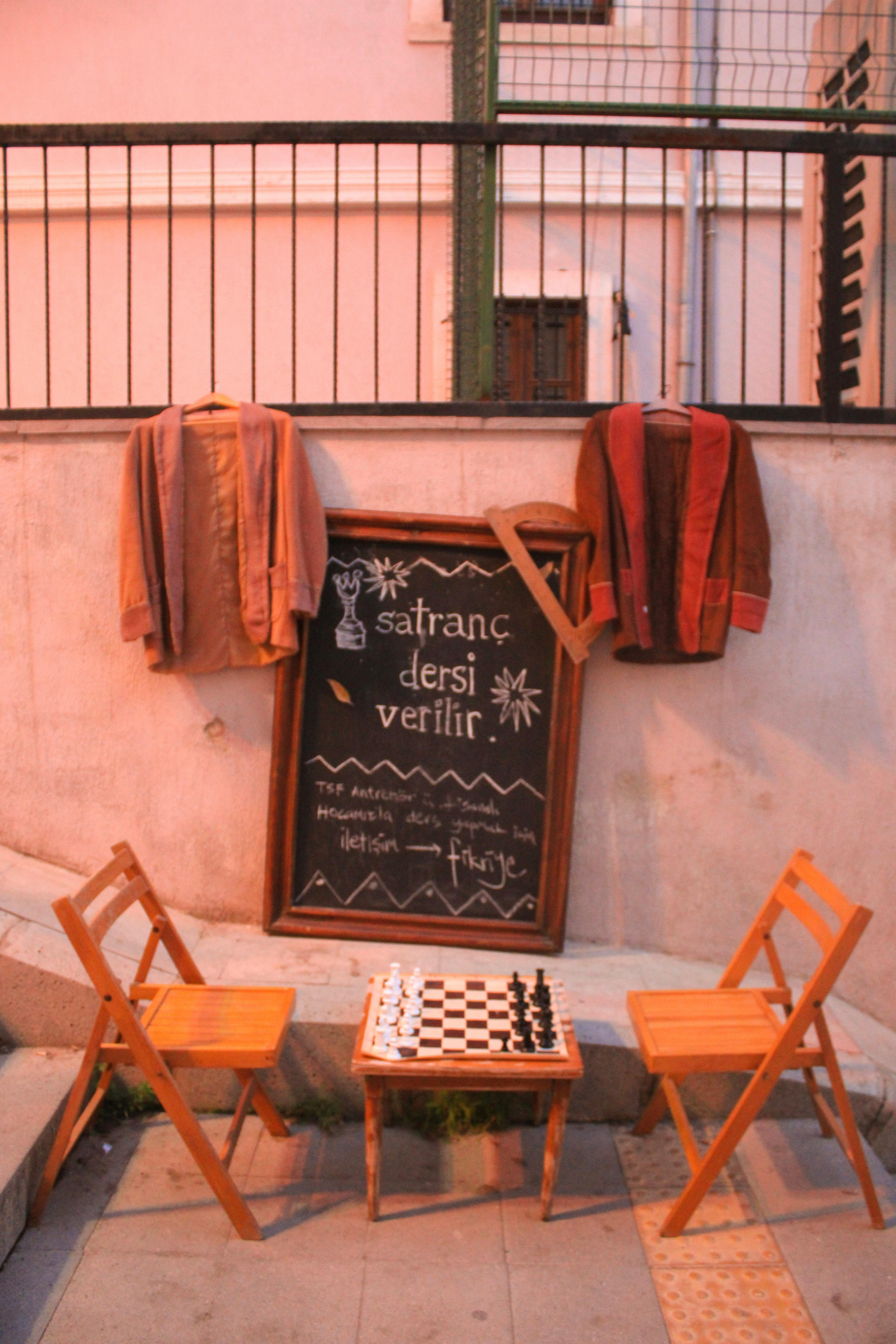 a chess board table with two chairs in front of a small blackboard with coats hanging on a railing on a stairway alley