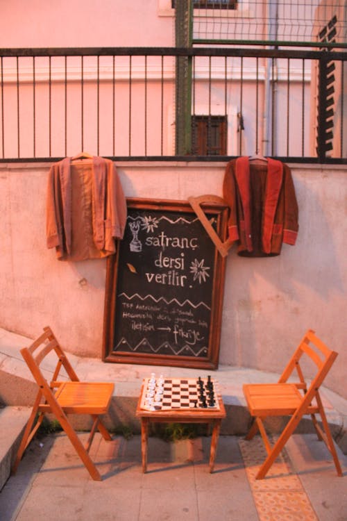 A Chess Board Table With Two Chairs In Front Of A Small Blackboard With Coats Hanging On A Railing On A Stairway Alley