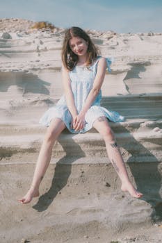 A young woman in a blue dress sits barefoot on sand, enjoying a sunny day outdoors.