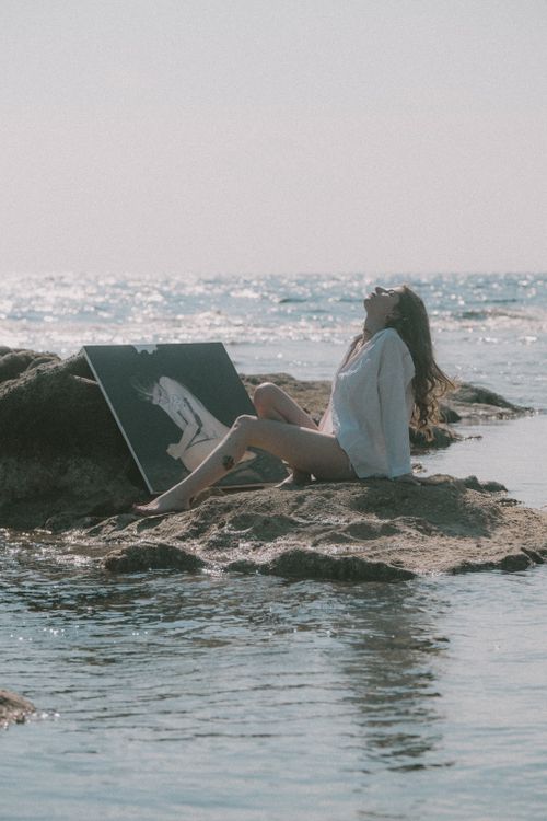 Free A serene woman sits by the ocean with a painting, exuding bohemian artistry against a summer backdrop. Stock Photo