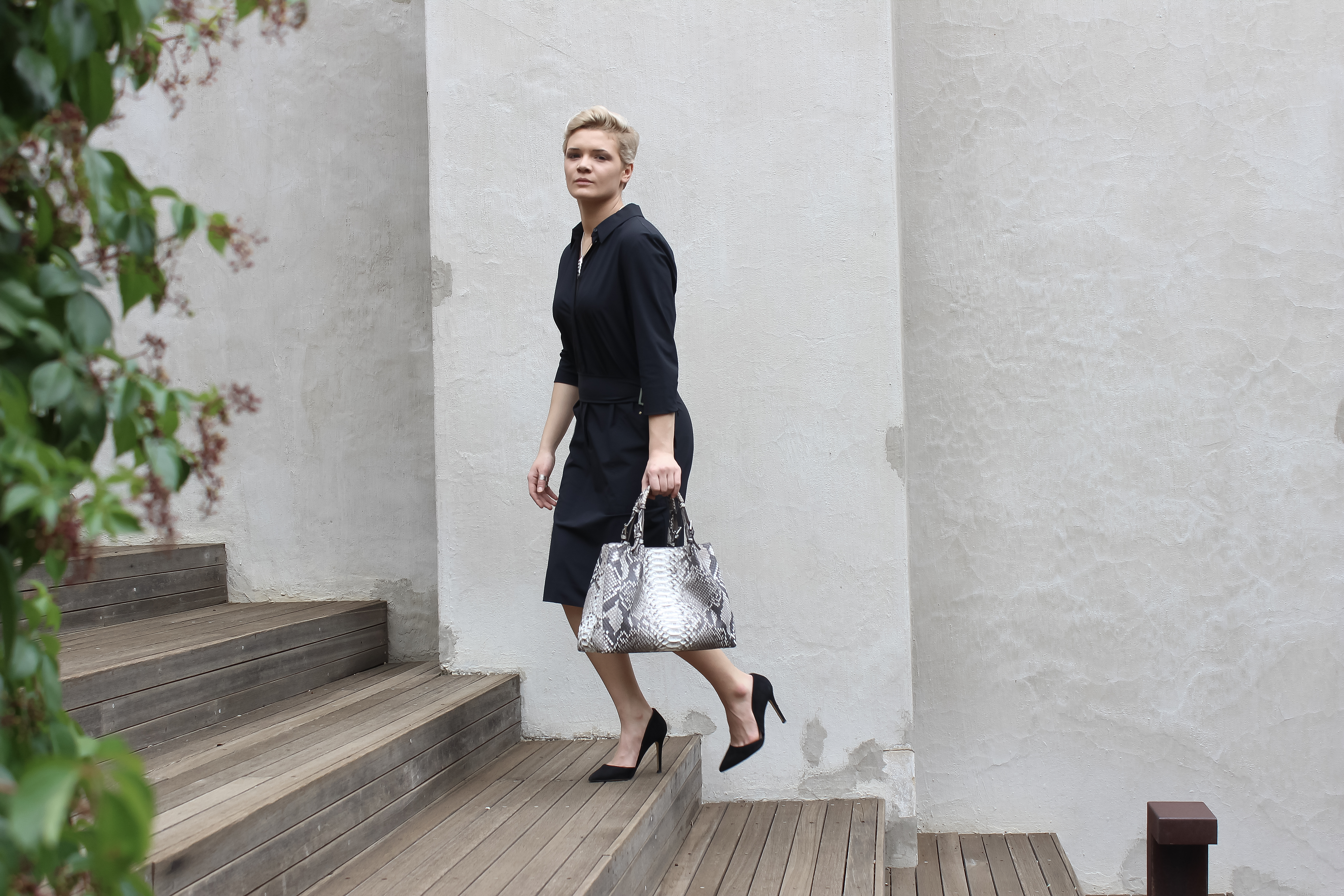 Elegant woman in a black dress with snakeskin handbag walking on wooden steps.