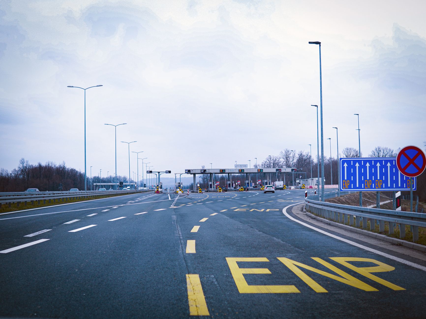 Wide highway leading to a toll plaza under a cloudy sky, featuring road signs and empty lanes.