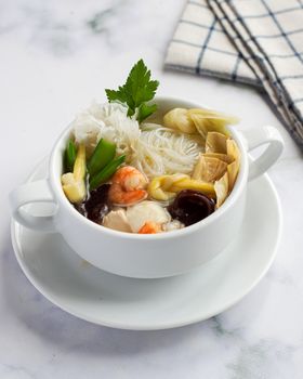 White bowl of Asian seafood soup with shrimp, mushrooms, and noodles on a white table. - Fotografer Sidoarjo