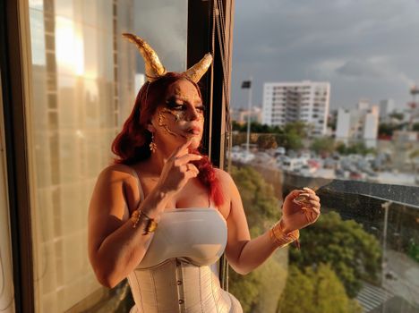 Woman in costume with gold makeup and horns, standing by a window at sunset.