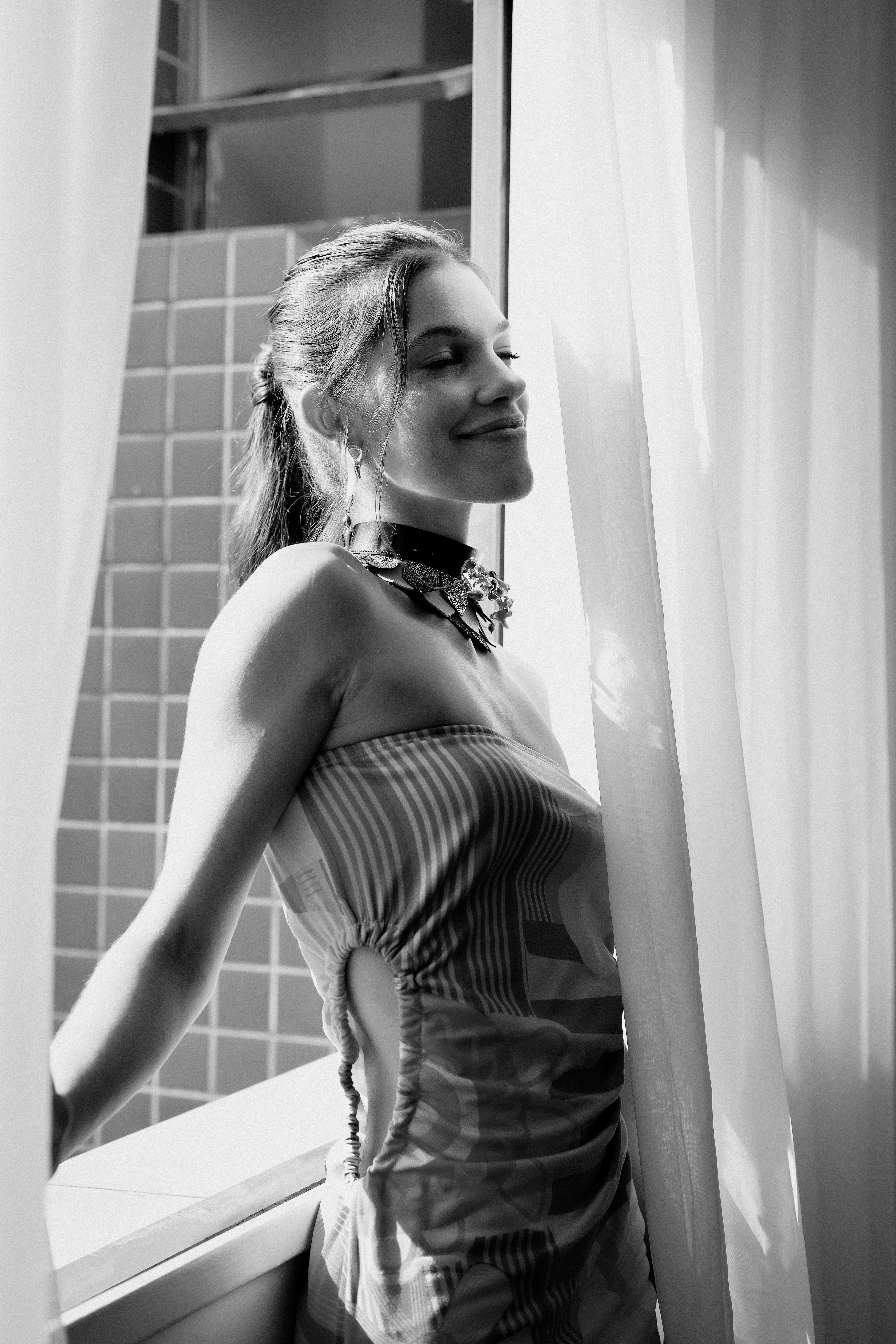 Stylish woman in elegant dress poses by window in a black and white photograph.