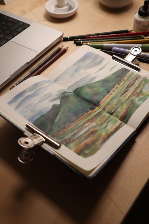 Free Top view of an open sketchbook with a landscape painting beside art supplies. Stock Photo