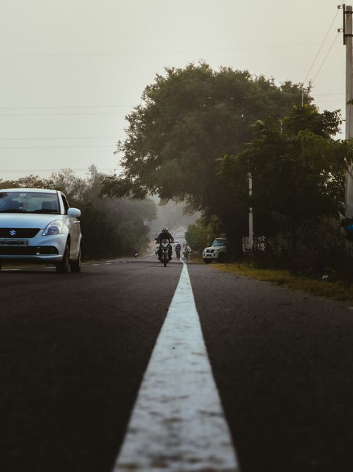 Free A tranquil rural road scene with cars and motorcycles surrounded by lush greenery and a warm dusk atmosphere. Stock Photo