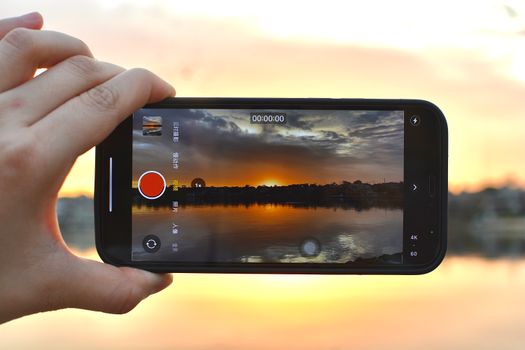 A person uses a smartphone to capture a vivid sunset by the lake, reflecting colors.
