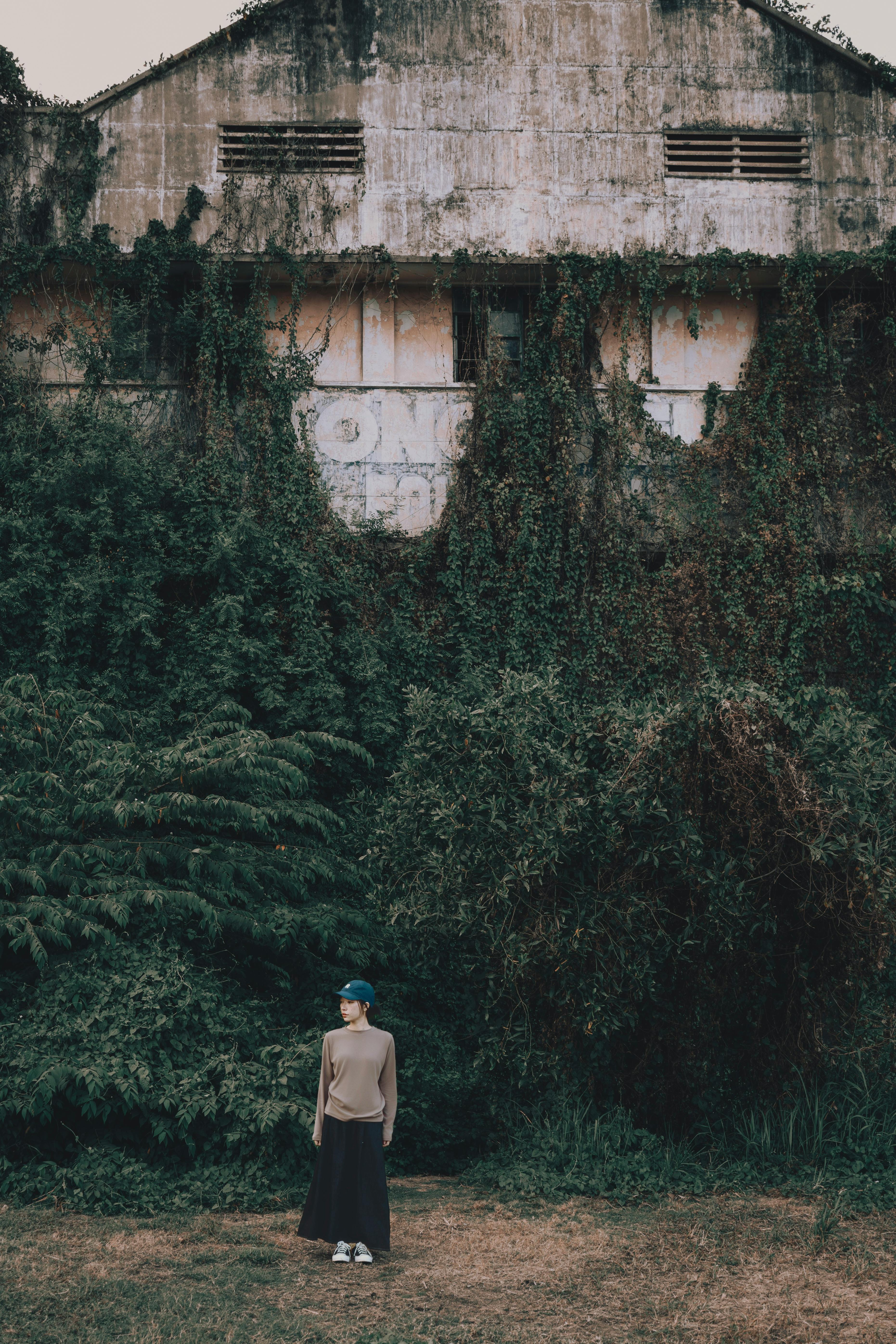 Person standing near an abandoned building covered in dense greenery.