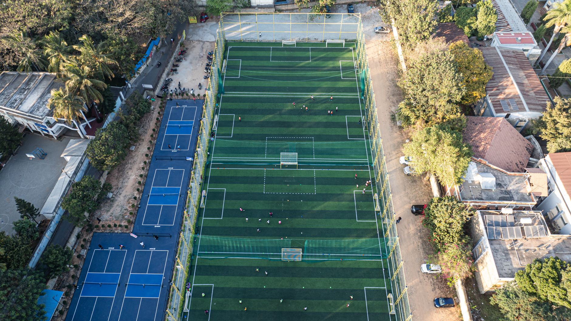 Aerial shot of soccer and tennis courts surrounded by buildings and trees, showcasing urban sports facilities.