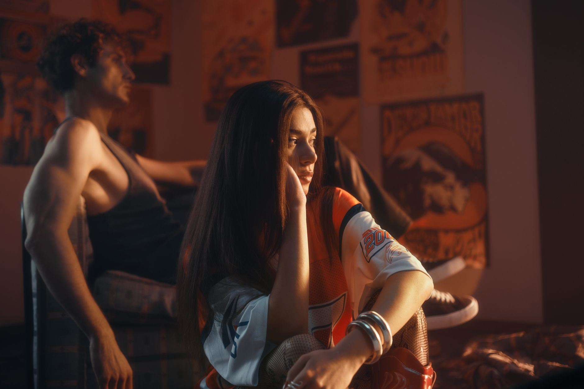 A young couple sits in a dimly lit room with vintage posters, creating a nostalgic and introspective atmosphere.