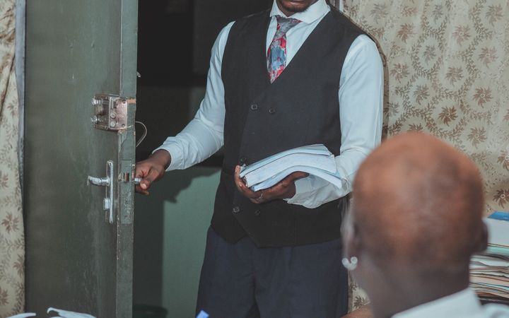 A young man in formal attire enters an office holding documents, depicting a professional setting.