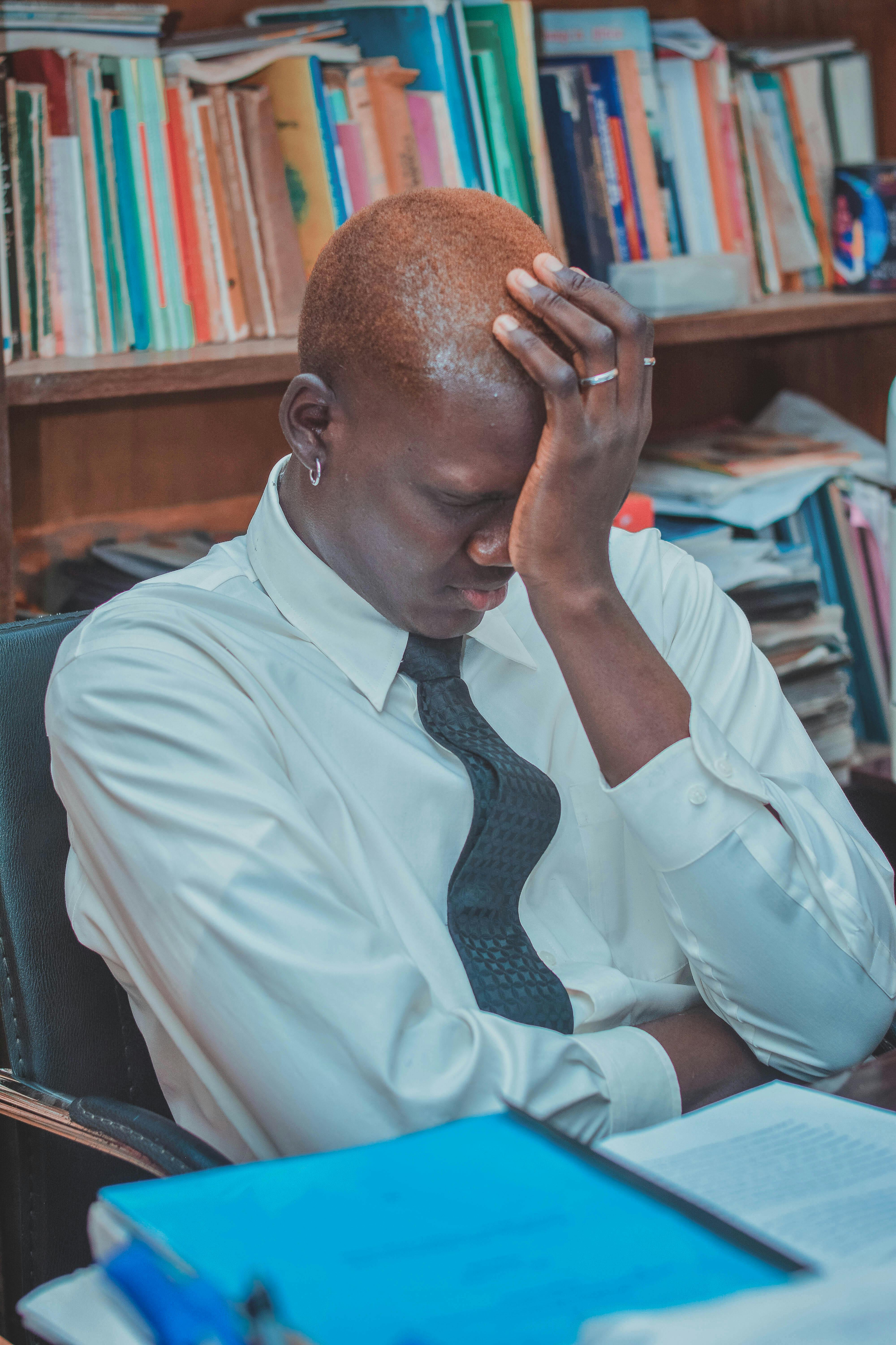 A tired businessman holds his head, seated at work amidst books.