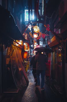 Two people with umbrellas walk through a brightly lit Tokyo alleyway at night, featuring neon lights and lanterns.