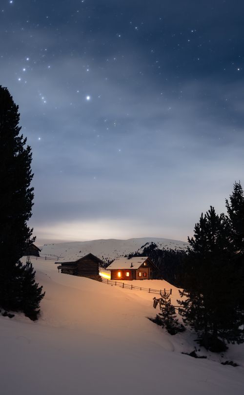 Free A warm-lit cabin nestled in snowy Italian mountains under a starlit sky. Stock Photo