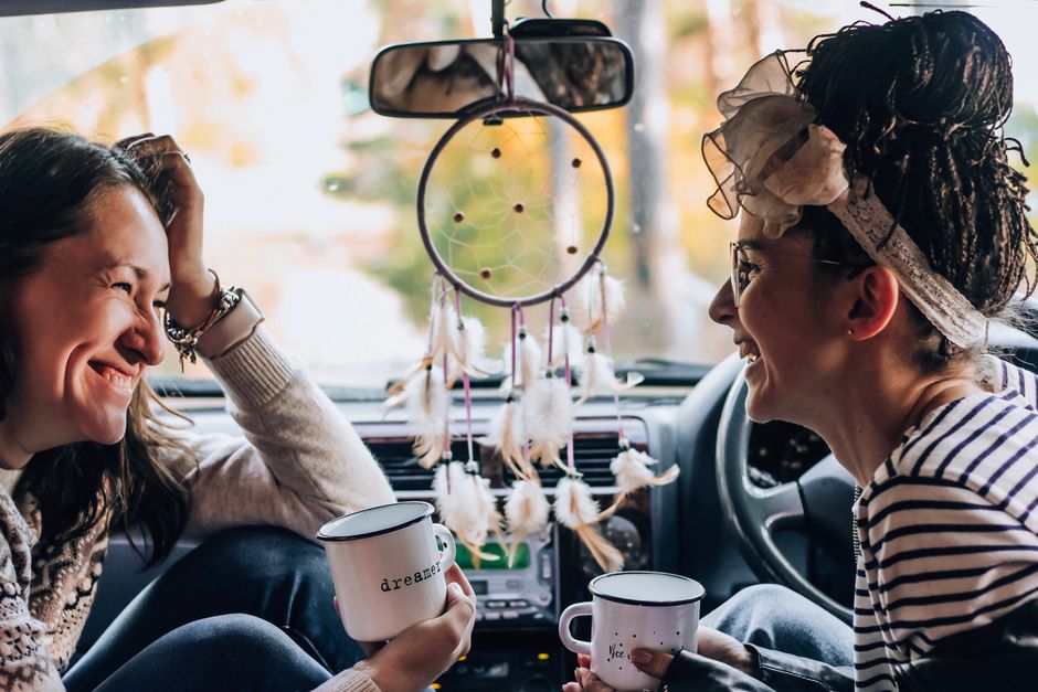 Two women enjoying a cheerful conversation in a parked car with mugs and a dreamcatcher.