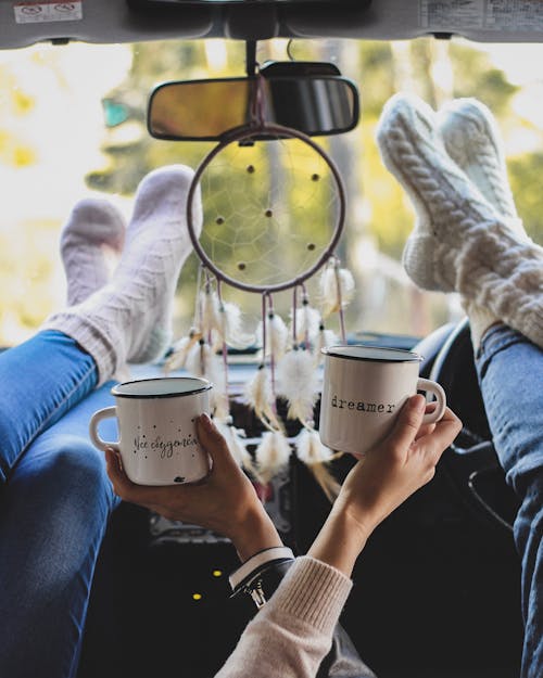 Deux Personnes Tenant Des Tasses à Café Avec Leurs Pieds Sur Le Tableau De Bord D'un Véhicule Avec Un Ornement Accroché Sur Le Rétroviseur