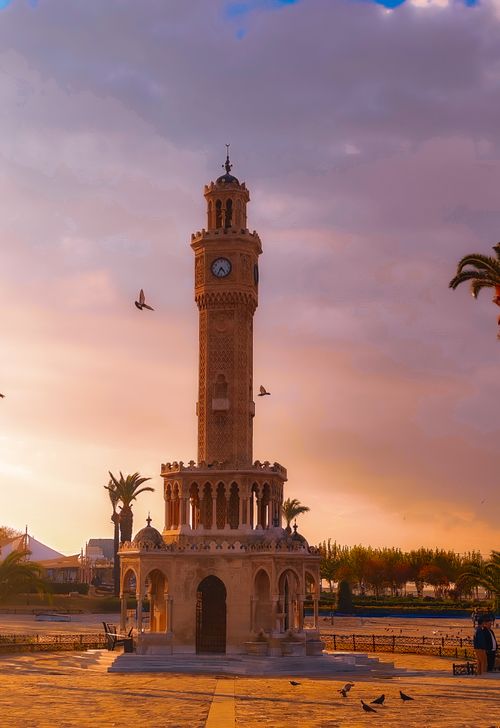 Free The historic Izmir Clock Tower stands under a vibrant sunset sky, framed by palms and birds. Stock Photo