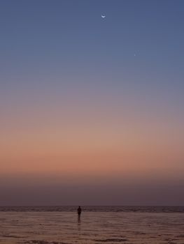A peaceful Indian beach at sunset with a crescent moon and lone figure in silhouette.