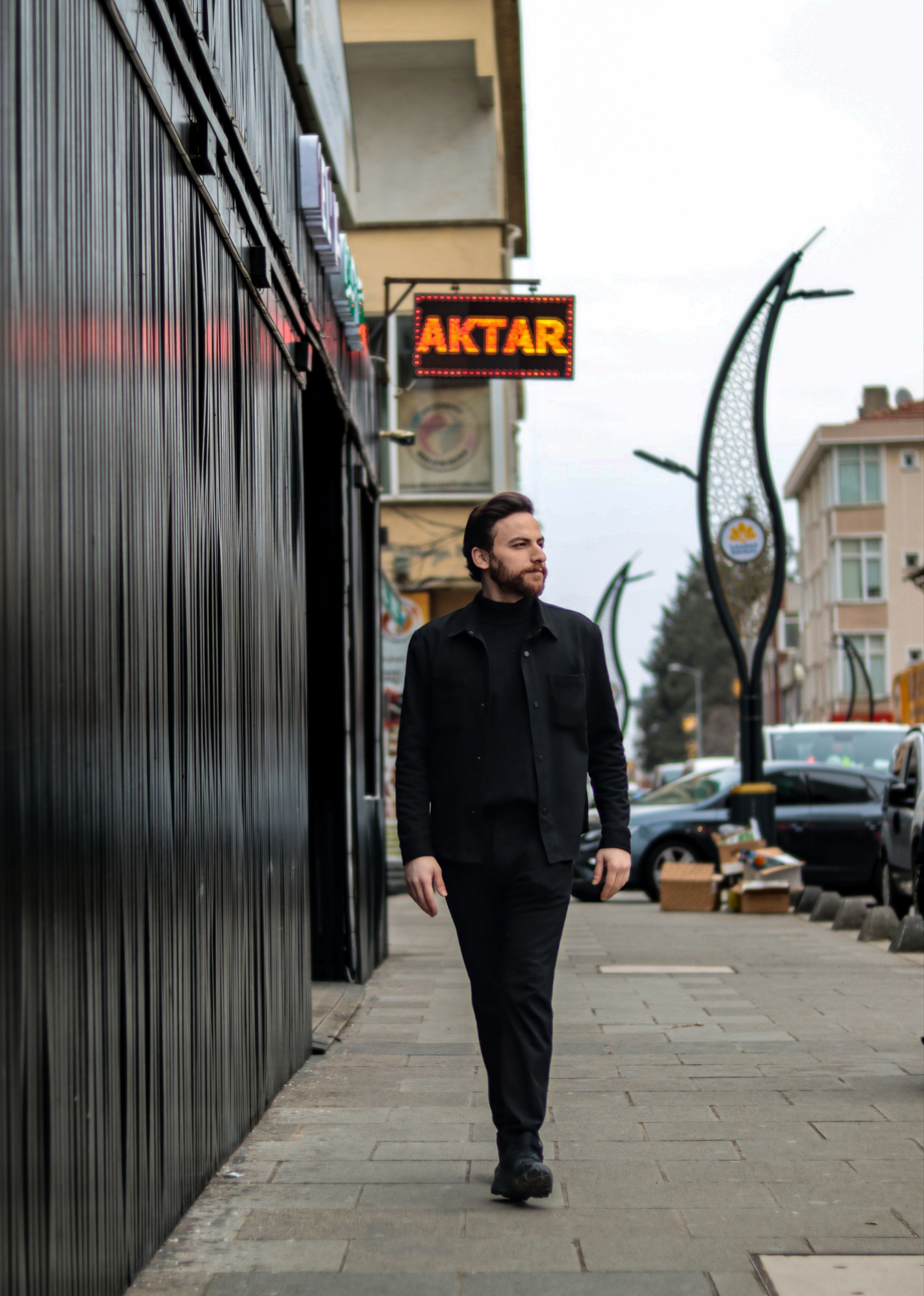 A man in black clothing walking down a city sidewalk past a neon sign reading 'AKTAR'.
