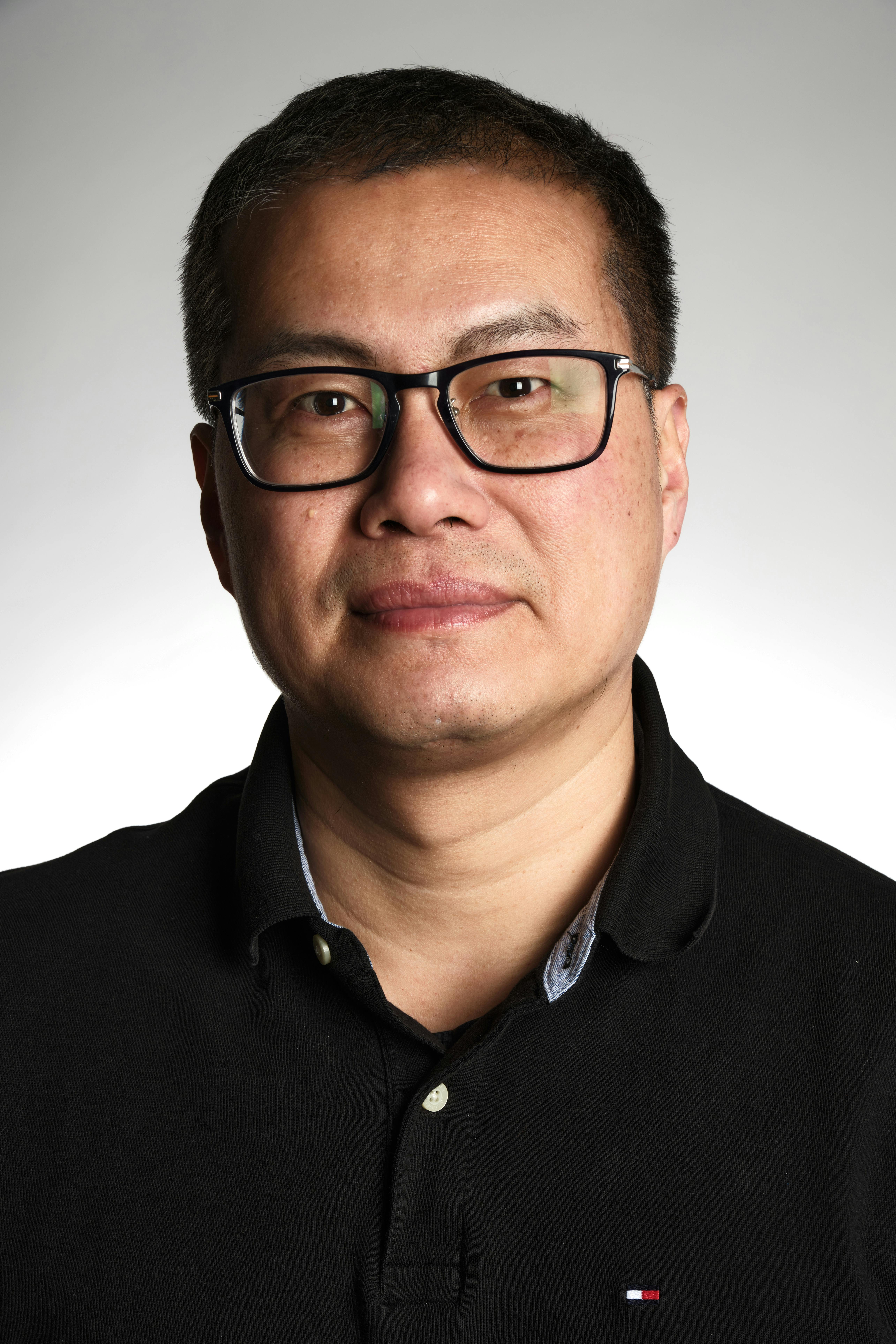 Close-up portrait of a man in glasses and black polo against a soft gradient background.