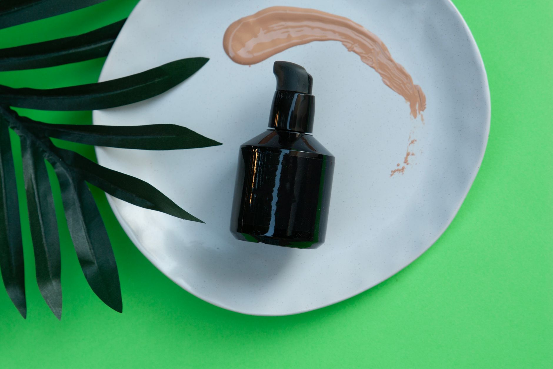 Black skincare pump bottle on a white plate with green leaf and makeup smear.