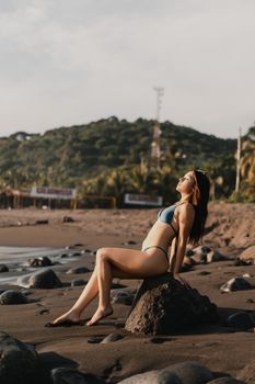 A woman in a bikini enjoys a sunny day on a rocky beach with lush scenery.