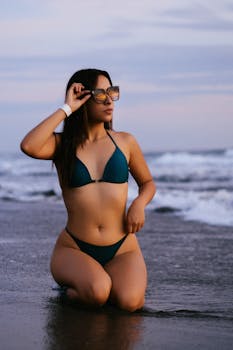 A woman in a bikini poses by the ocean during sunset, exuding confidence and style.