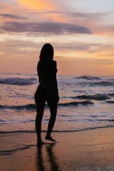 A silhouette of a woman walking along the beach as the sun sets over the ocean waves.
