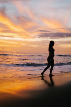 A woman walks along the beach during a vibrant sunset, creating a serene silhouette.