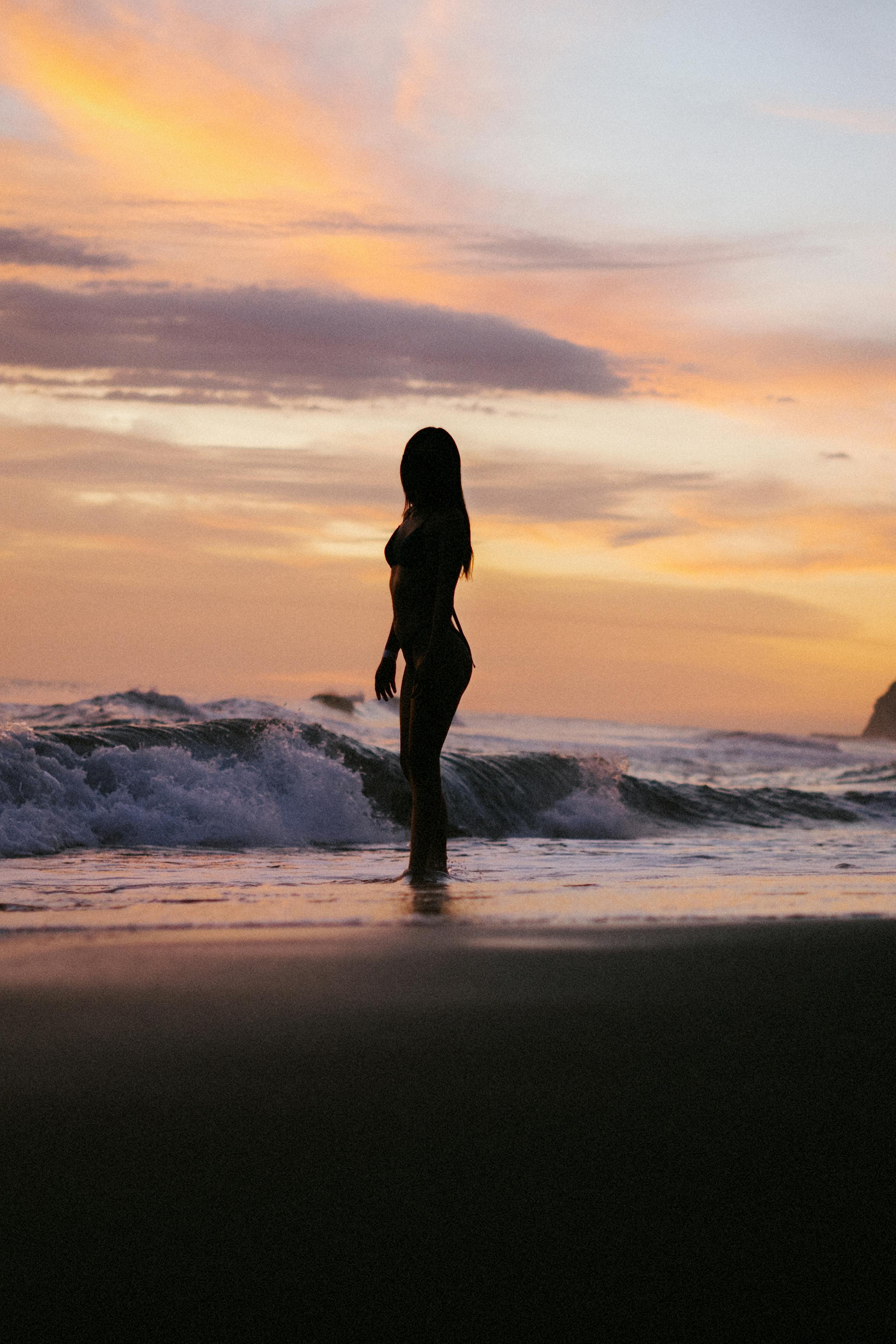 A serene silhouette of a woman standing on a beach as waves crash at sunset.