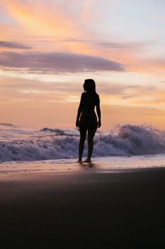 A woman in silhouette walks along a beach during a captivating sunset, embraced by waves.