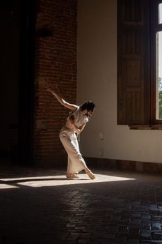 Woman in a modern dance pose in a sunlit historic room with brick walls.