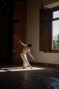 Dancer elegantly poses in sunlit room with rustic brick wall and open window.