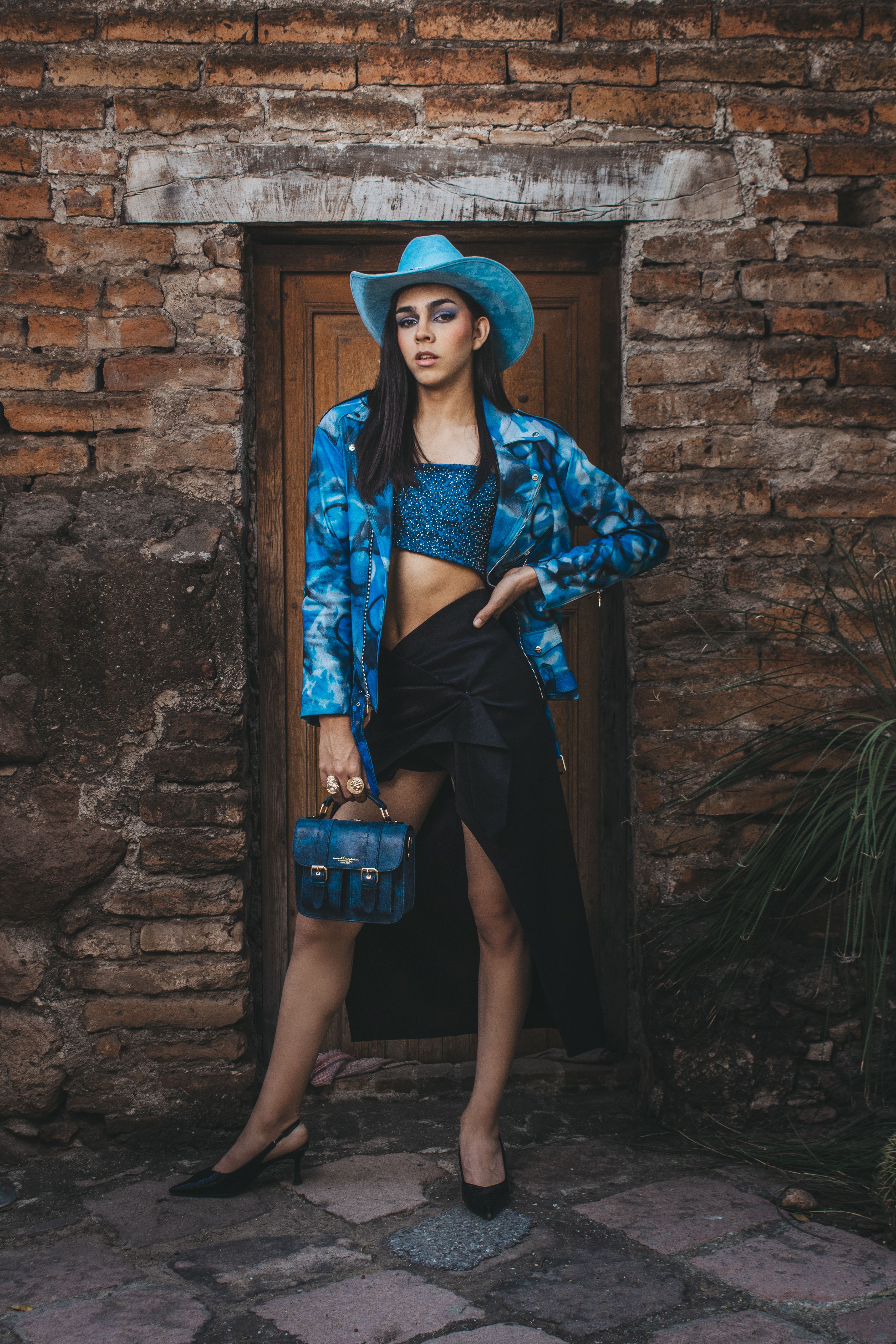Stylish woman in blue attire and cowboy hat posing outdoors against rustic brick wall.