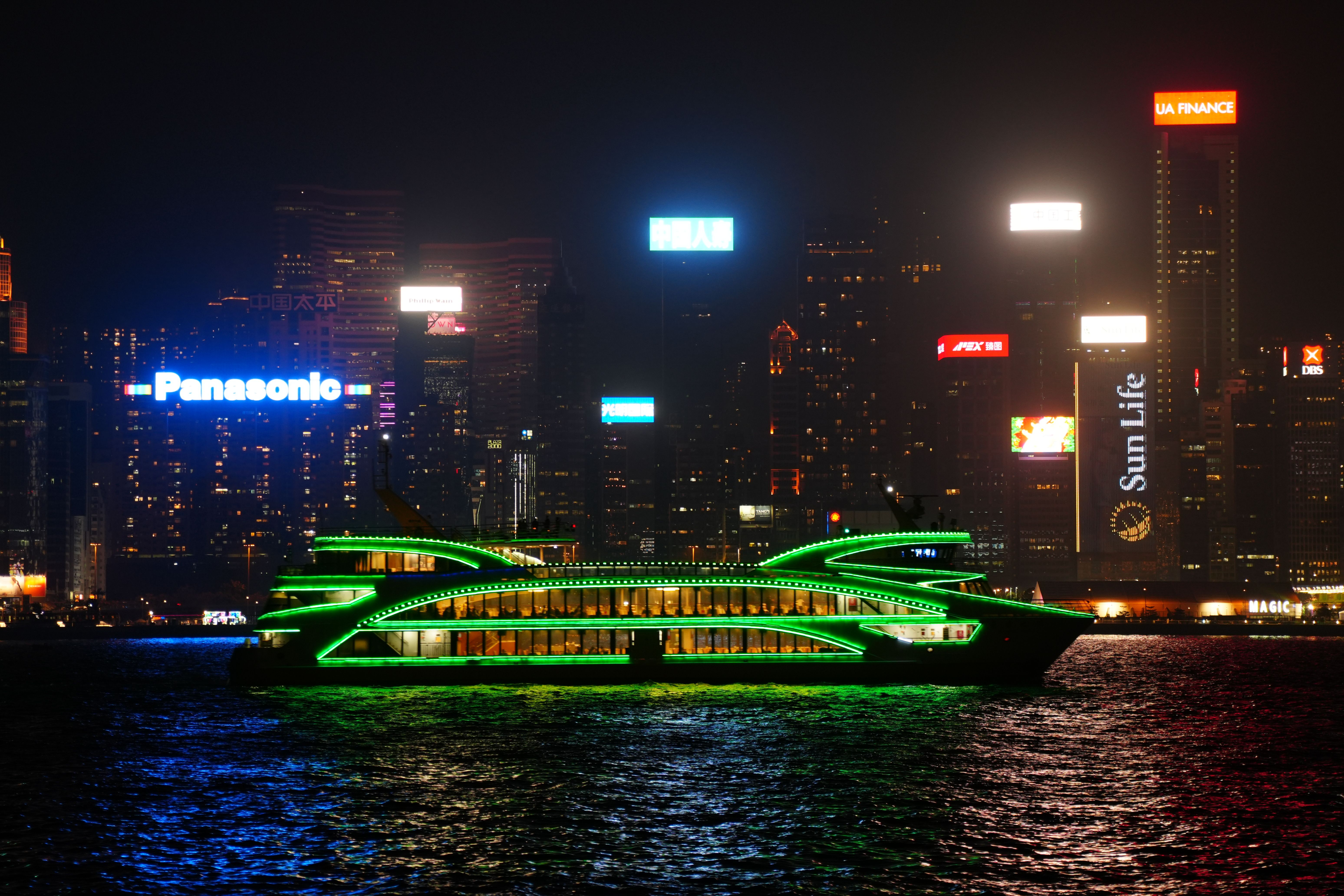 A neon-lit ferry cruises in front of the vibrant Hong Kong cityscape at night.