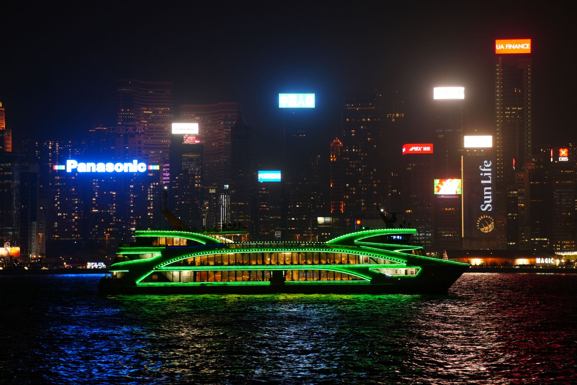 A neon-lit ferry cruises in front of the vibrant Hong Kong cityscape at night.