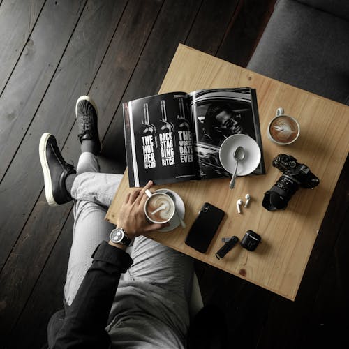 Вид сверху на стол с мужчиной, держащим чашку латте с журналом, камерой, мобильным телефоном и другими личными вещами